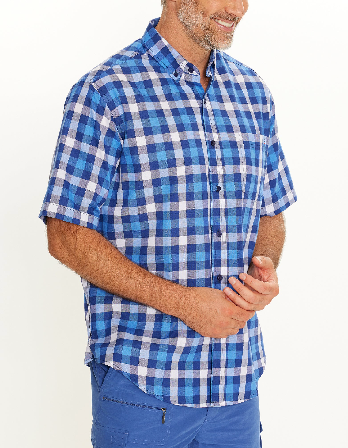 A man wearing the Breakaway Gil Oxford Shirt, a comfortable short-sleeve button-up with a blue and white plaid design, paired with blue pants. He is smiling while standing against a plain white background.