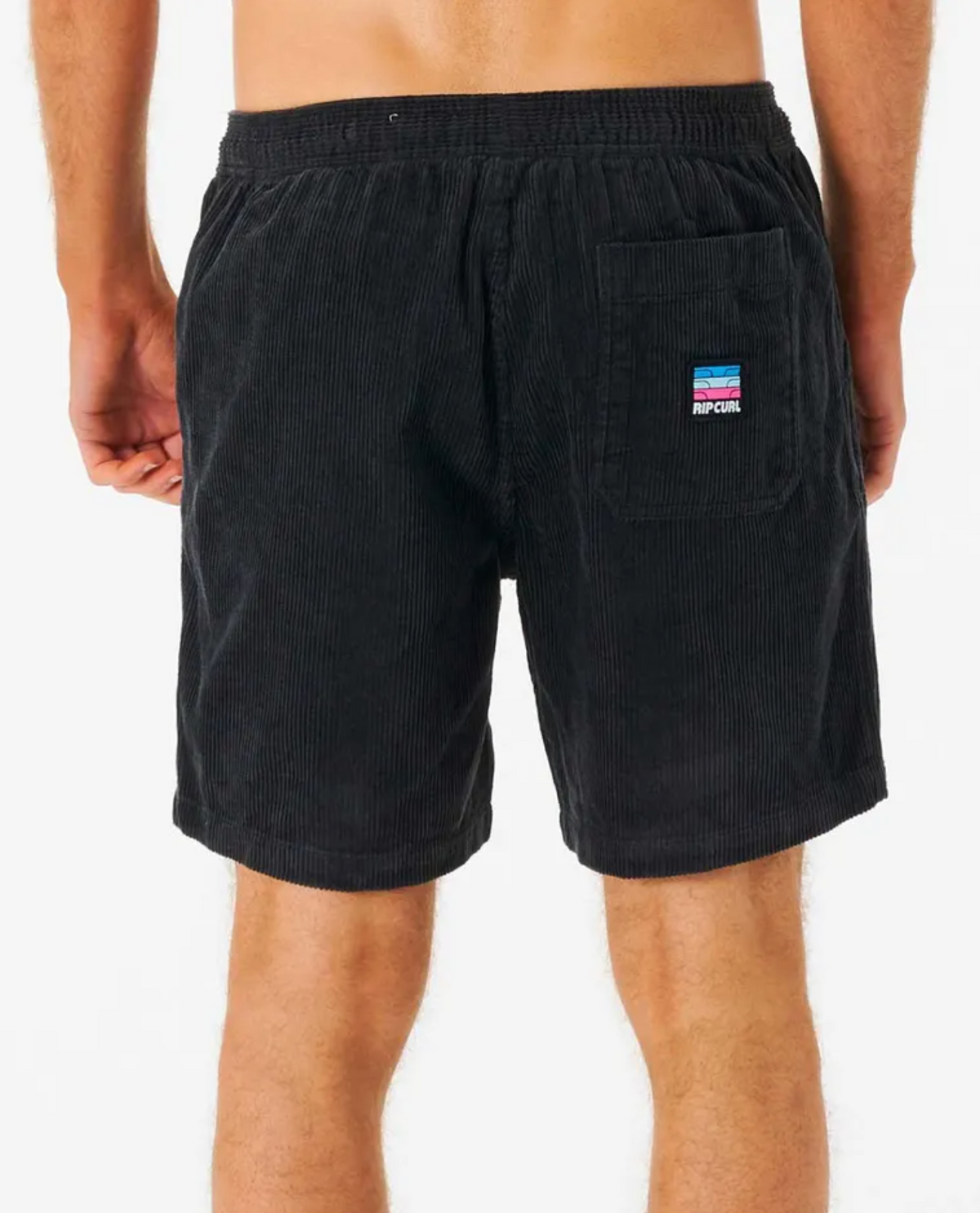 A person models the Rip Curl Surf Revival Cord Volley, featuring black cotton corduroy shorts with an elastic waistband and a 90's baggy fit. The back pocket showcases a colorful rectangular patch with blue, pink, and white stripes against a white background.