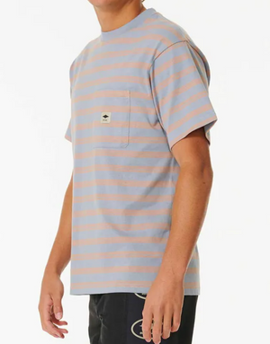 A person wears a Rip Curl Quality Surf Products Stripe Tee in light blue and beige with a chest pocket and the brand's square logo, against a white backdrop. The tee is paired with black pants featuring a circular design.