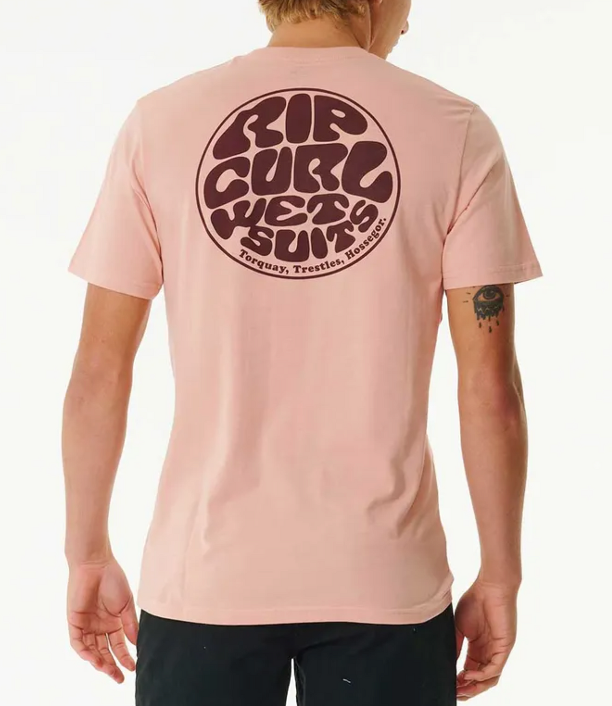 A person faces away, wearing the Rip Curl Wetsuit Icon Tee in light pink—a 100% cotton casual shirt with a large circular "Rip Curl Wetsuits" logo on the back and small text featuring Torquay, Trestles, and Hossegor.