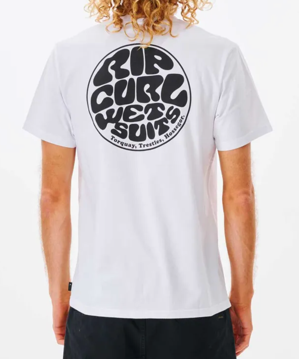 A person with curly hair is seen wearing the Rip Curl Wetsuit Icon Tee, a casual 100% cotton white shirt with a black circular graphic reading "Rip Curl Wetsuits" and "Torquay, Trestles, Hossegor" on the back. Facing away from the camera, they're stylishly paired with black pants.