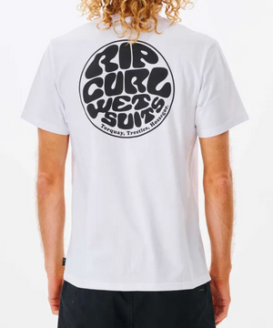 A person with curly hair is seen wearing the Rip Curl Wetsuit Icon Tee, a casual 100% cotton white shirt with a black circular graphic reading "Rip Curl Wetsuits" and "Torquay, Trestles, Hossegor" on the back. Facing away from the camera, they're stylishly paired with black pants.