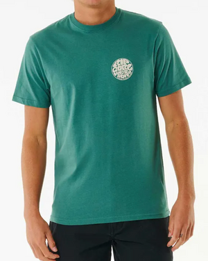 A person wears a comfortable teal Rip Curl Wetsuit Icon Tee, made from 100% cotton, featuring a small circular graphic logo with stylized text on the left chest. Paired with black pants, they stand against a plain white background.