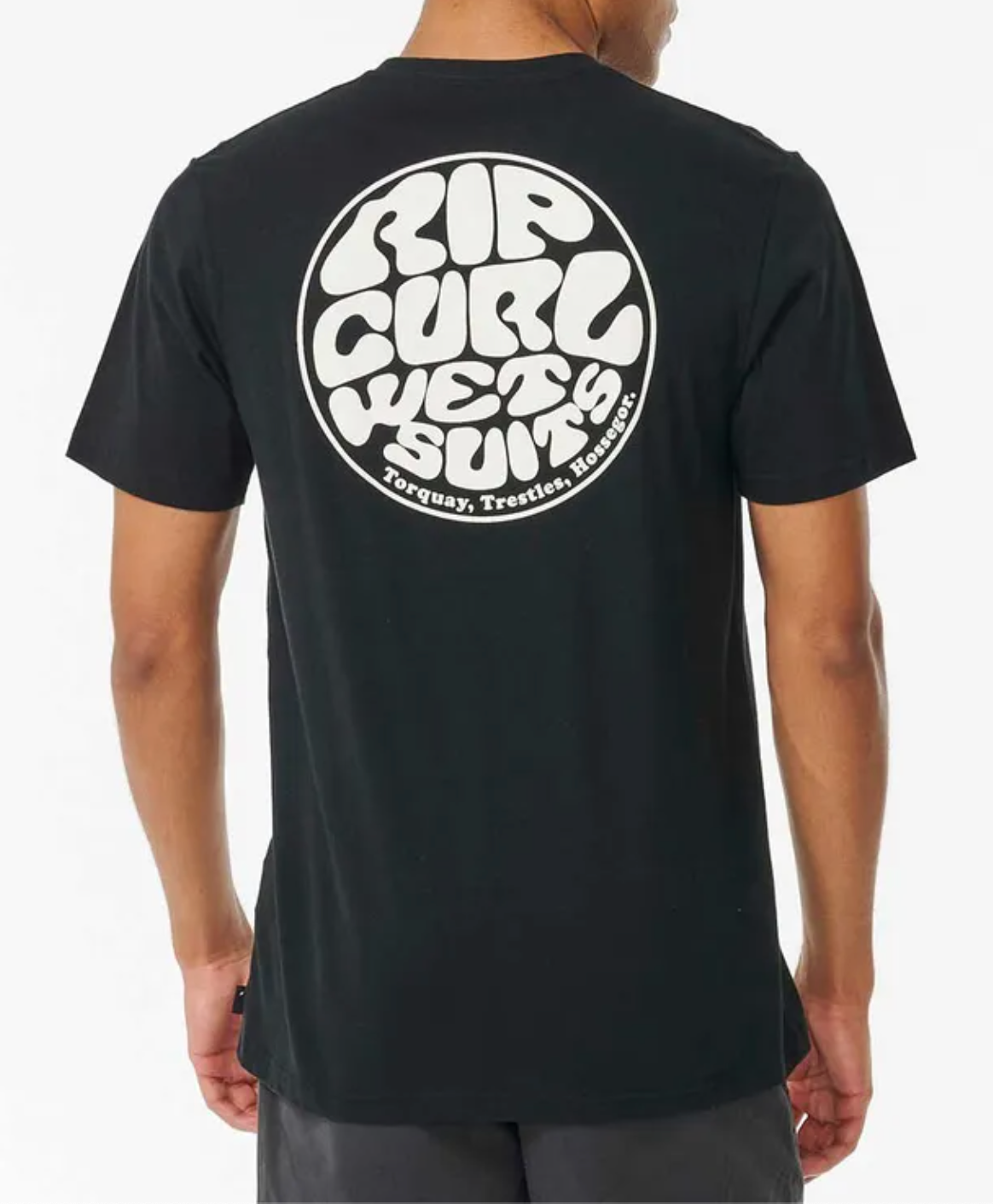 Man wearing the Rip Curl Wetsuit Icon Tee—black with a bold white circular "Rip Curl Wetsuits, Torquay, Trestles, Hossegor" graphic on the back. Made from 100% cotton for all-day comfort and casual style.