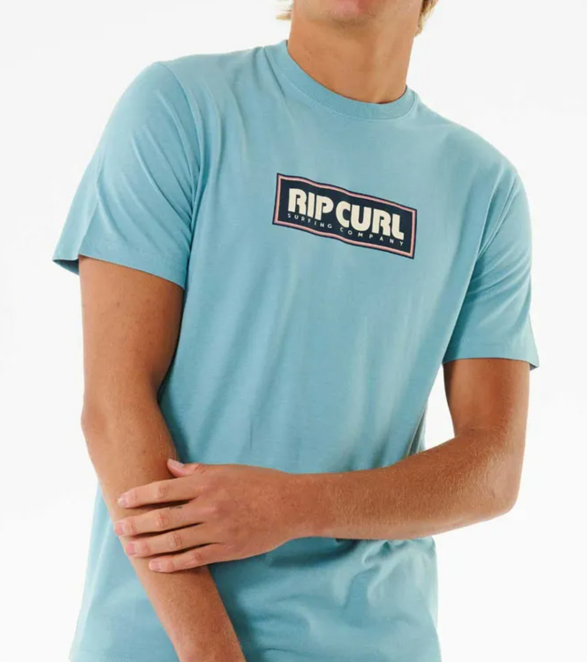 A person models the Rip Curl Big Mumma Icon Tee in light blue, made from organic cotton, standing against a plain background with one arm bent and hand touching the opposite forearm.
