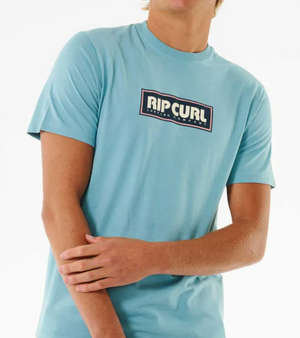 A person models the Rip Curl Big Mumma Icon Tee in light blue, made from organic cotton, standing against a plain background with one arm bent and hand touching the opposite forearm.
