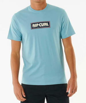 A person wears the Rip Curl Big Mumma Icon Tee in light blue, crafted from soft organic cotton with a rectangular chest logo, paired with black pants and standing against a plain white background.