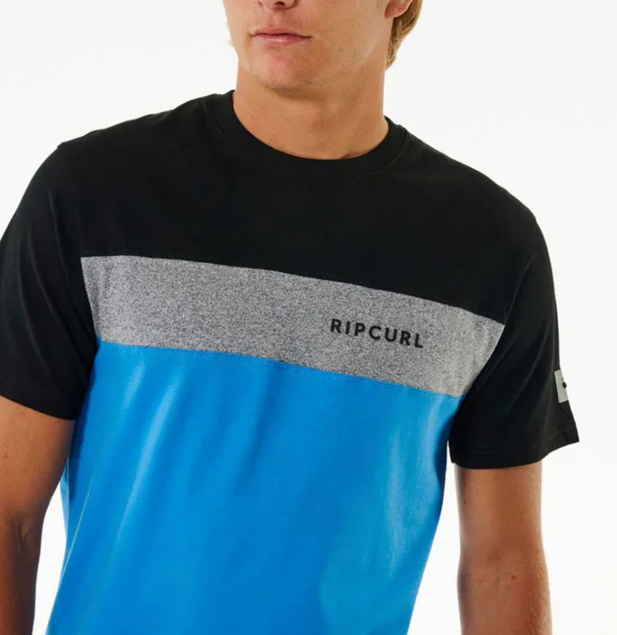 A person wearing the Rip Curl Undertow Panel Tee, a 100% cotton shirt with a black top, gray stripe with "RIP CURL" print, and bright blue lower half—blending style and comfort seamlessly.