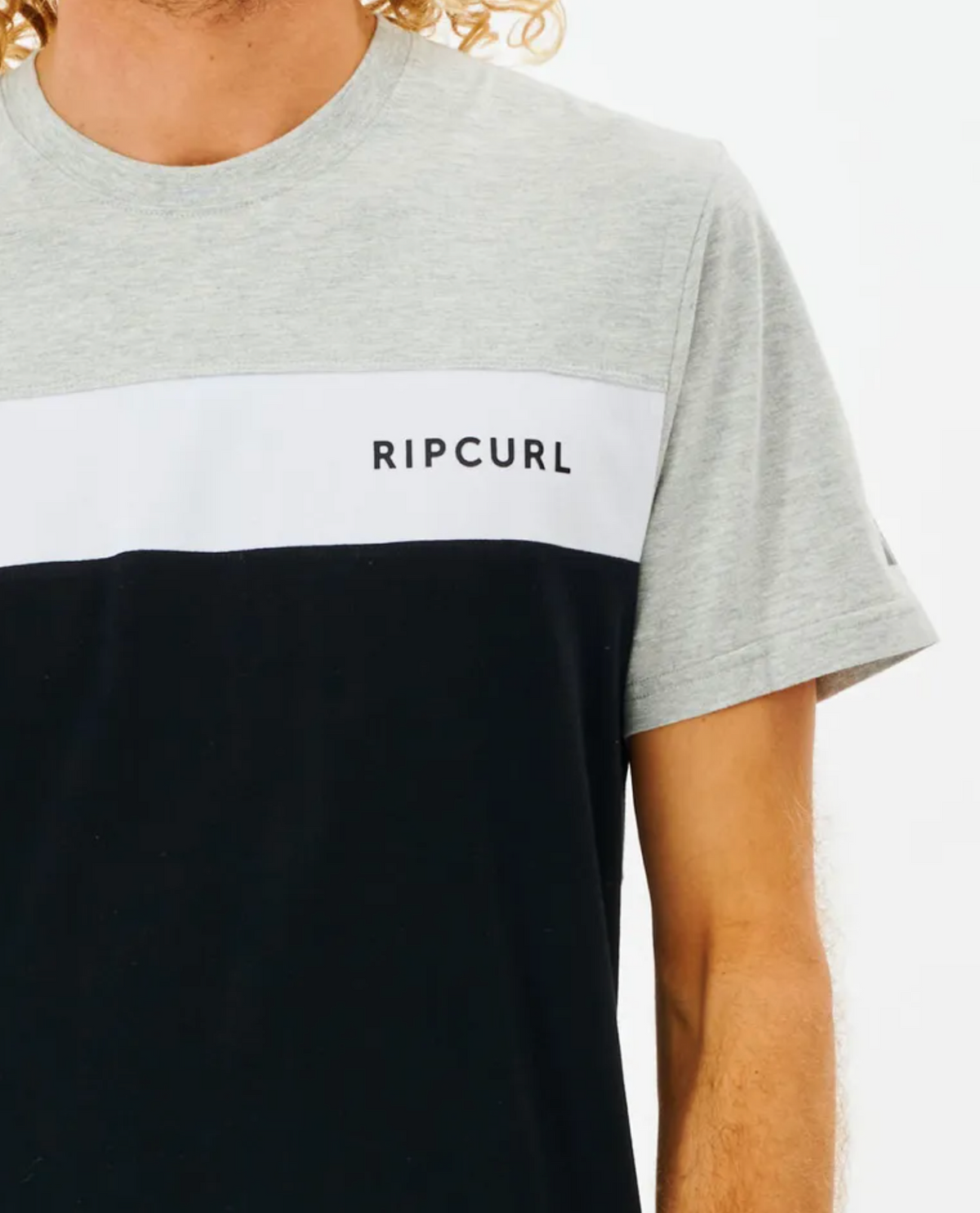A person is wearing the Rip Curl Undertow Panel Tee with a gray top, white stripe displaying "RIPCURL" in black letters, and a black bottom. Made from 100% cotton, this tee not only suits their curly hair but also offers unmatched comfort and style.