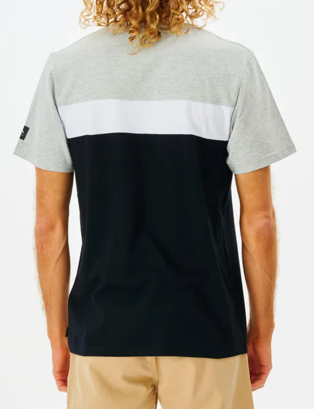 A person with curly hair is wearing a Rip Curl Undertow Panel Tee, featuring gray, white, and black horizontal sections. This 100% cotton tee pairs well with their beige pants as they face away from the camera.