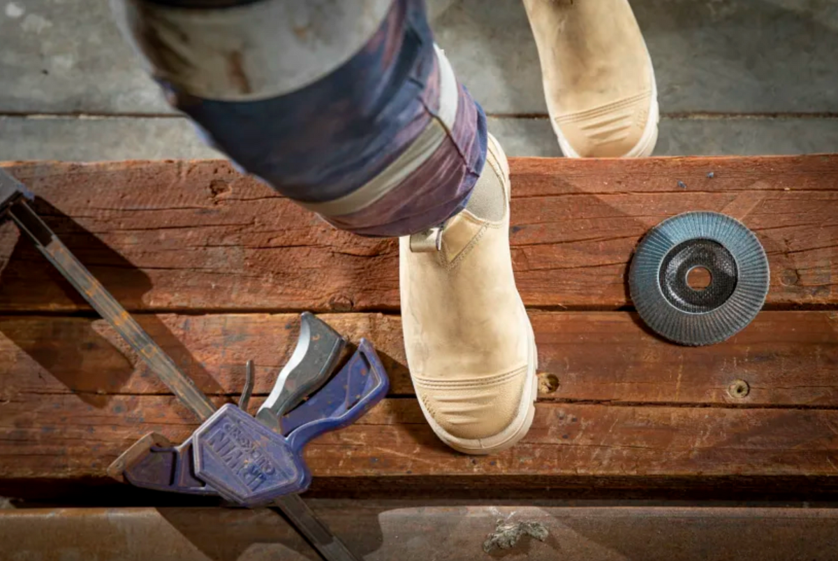 A worker wearing Steel Blue Hobart Scuff Cap Elastic Sided Safety Boots with a 200J steel toe cap steps on a wooden surface, with a purple clamp and a circular saw blade nearby. Rails and screws are also visible in the industrial working environment.