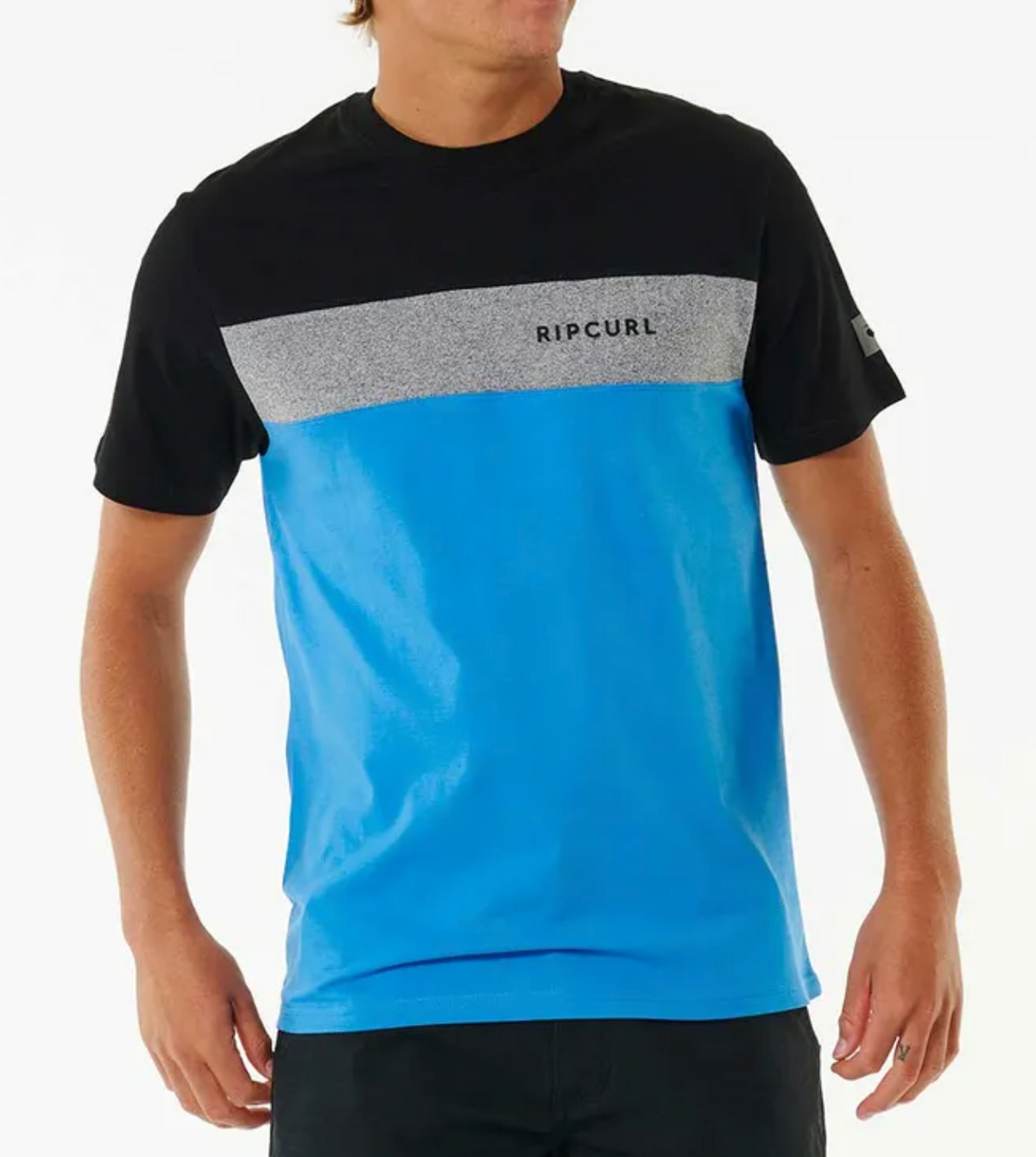 A person wears the Rip Curl Undertow Panel Tee, a 100% cotton shirt with a black upper part, gray stripe with "Rip Curl" text across the chest, and bright blue lower section. The individual stands with hands at their sides.