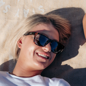 A person with short blond hair and a white shirt is lying on a light surface, smiling in the sunlight while wearing SafeStyle Fusions Brown Tort Frame Polarised Lens sunglasses.