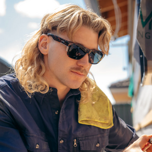 A man with long blonde hair and a mustache wears SafeStyle Fusions Brown Tort Frame Polarised Lens sunglasses and a navy shirt, with a yellow towel over his shoulder, sitting outdoors on a sunny day.