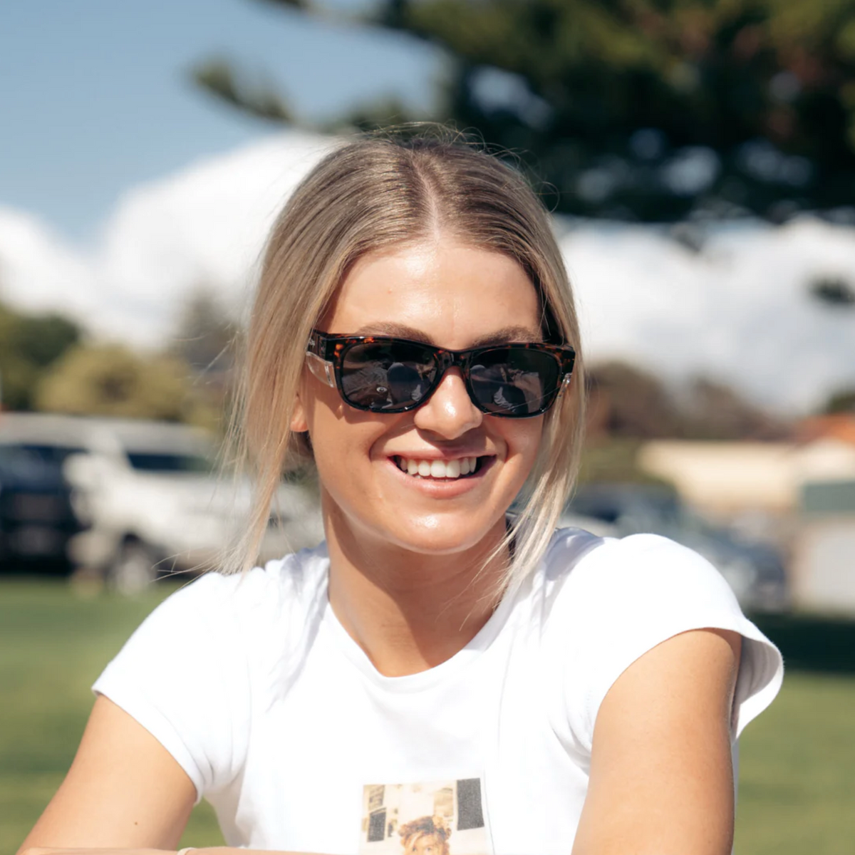 A smiling person with long blonde hair, wearing SafeStyle Classics Brown Tort Frame Polarised Lens sunglasses and a white t-shirt, sits outdoors on a sunny day. Green grass, tall trees, and cloudy skies highlight the importance of UV protection in eyewear.