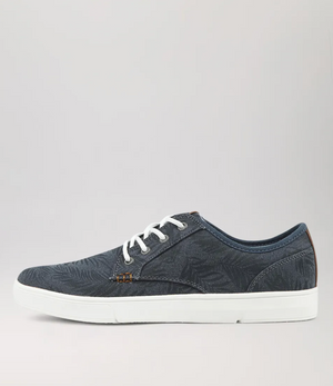 The Colorado Albatross Canvas Sneaker in dark blue showcases subtle leaf patterns and white laces, offering a wide fit for comfort. Its white rubber sole and memory foam inner ensure a cozy step. The shoe is pictured from the side on a plain gray background.