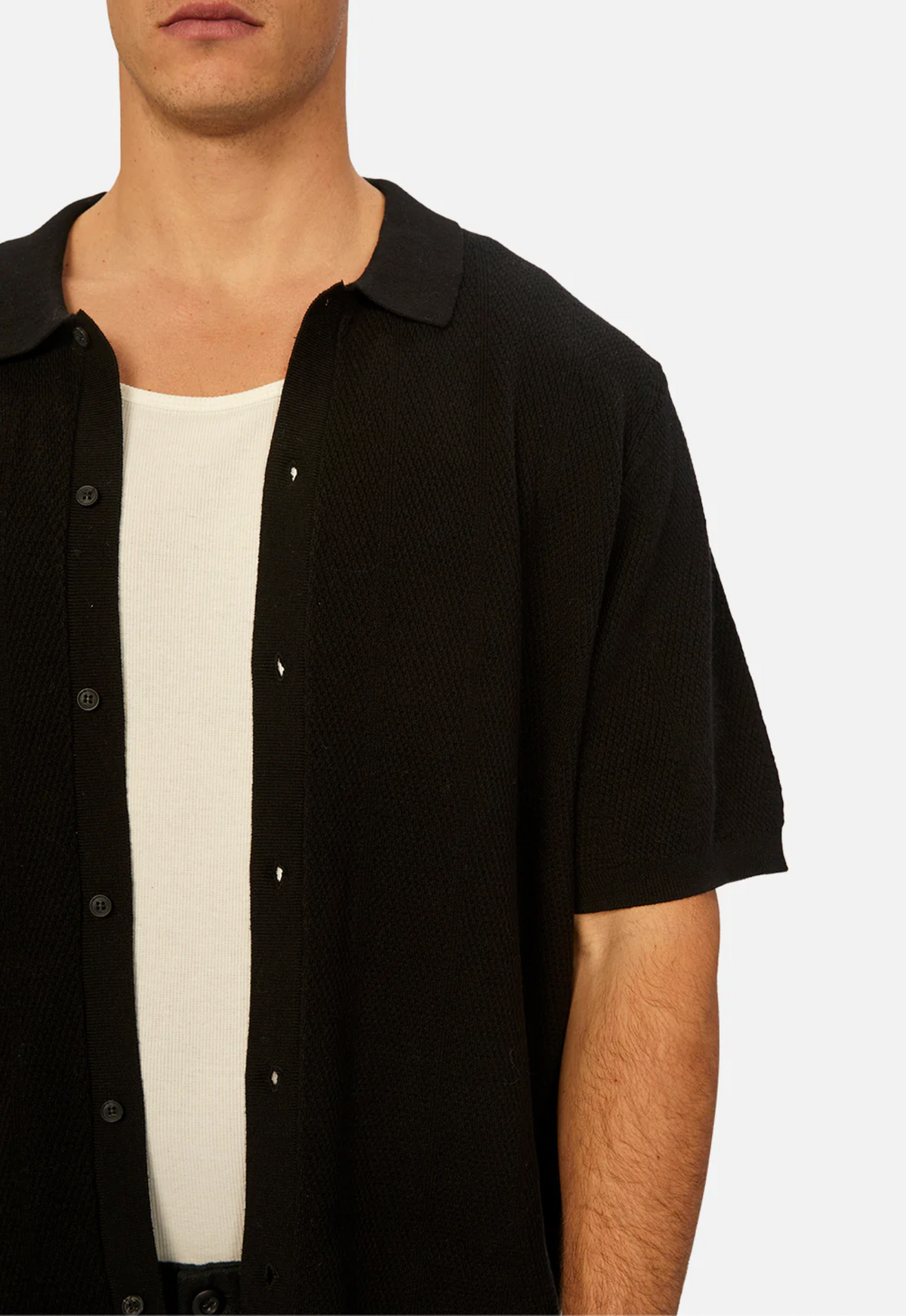 A man in an unbuttoned black Industrie The Modena Short Sleeve Shirt, made of 100% cotton, is layered over a white tank top against a plain backdrop, creating a relaxed and casual look with only the upper half of his body visible.