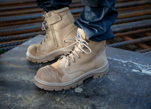 A person in Steel Blue Argyle Zip Scuff Cap Boots with a safety steel toe and blue jeans stands on metal at a construction site, with steel rebar in the background.