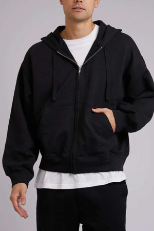 An individual is dressed in a black Silent Theory Zip Hoodie, composed of 80% cotton and 20% polyester, layered over a white t-shirt. Their left hand rests comfortably in the stylish hoodie's pocket against a plain grey background.