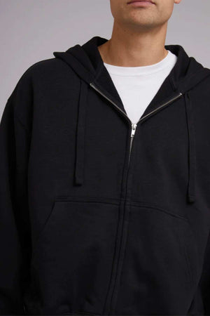 A person sports a chic and comfortable Silent Theory Zip Hoodie in black over a white t-shirt. Made of 80% cotton and 20% polyester, the hoodie features a front pocket and drawstrings dangling from the hood. Only the lower part of their face and upper chest are visible against a gray background.