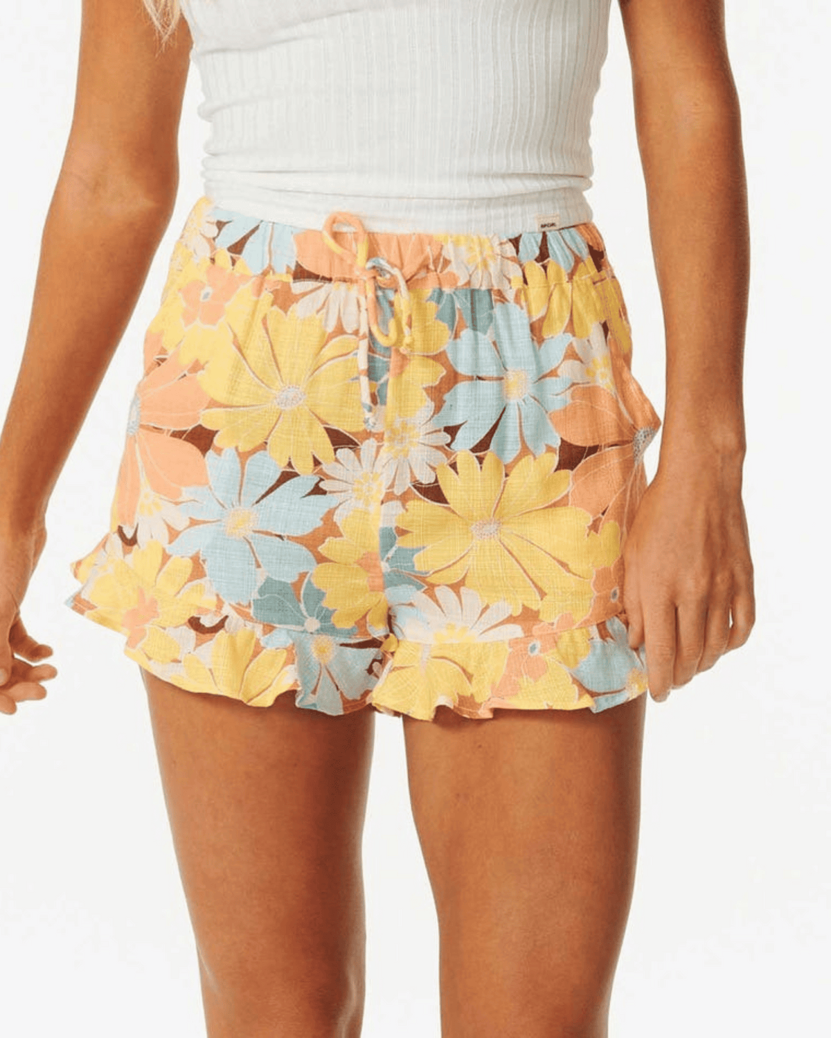 A person wearing a white ribbed tank and Rip Curl Sunrise Session Short, which features a yellow, peach, and blue print with ruffled trim and a drawstring waistband; image cropped to the torso and upper legs.