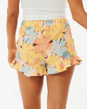 A person wears the Rip Curl Sunrise Session Short, a high-waisted Ecovero ruffled short with a colorful print, paired with a white ribbed tank top. The photo is taken from the back to showcase the shorts' design.