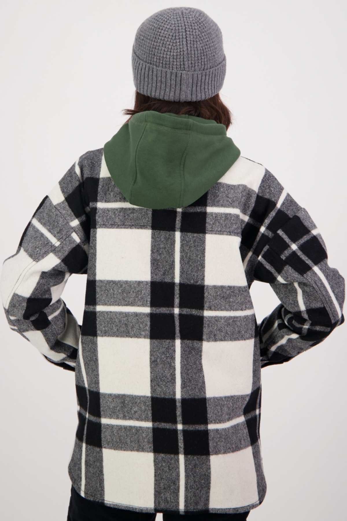 A person with long hair is wearing a gray knit beanie and a black-and-white checkered Swanndri Anchorage v2 Wool Shirt Jacket with a green hood. They are standing with their back to the camera, hands on their hips.