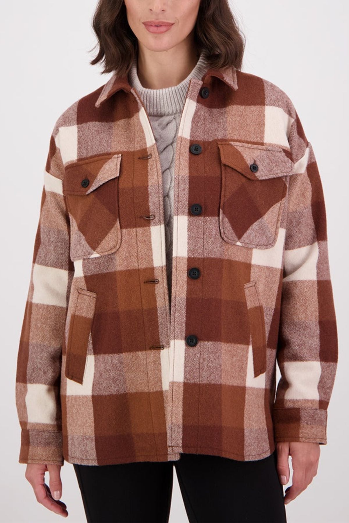 A woman wears the Swanndri Anchorage v2 Wool Shirt Jacket in brown and cream check over a light gray cable knit sweater, showcasing its front flap pockets and stylish design.