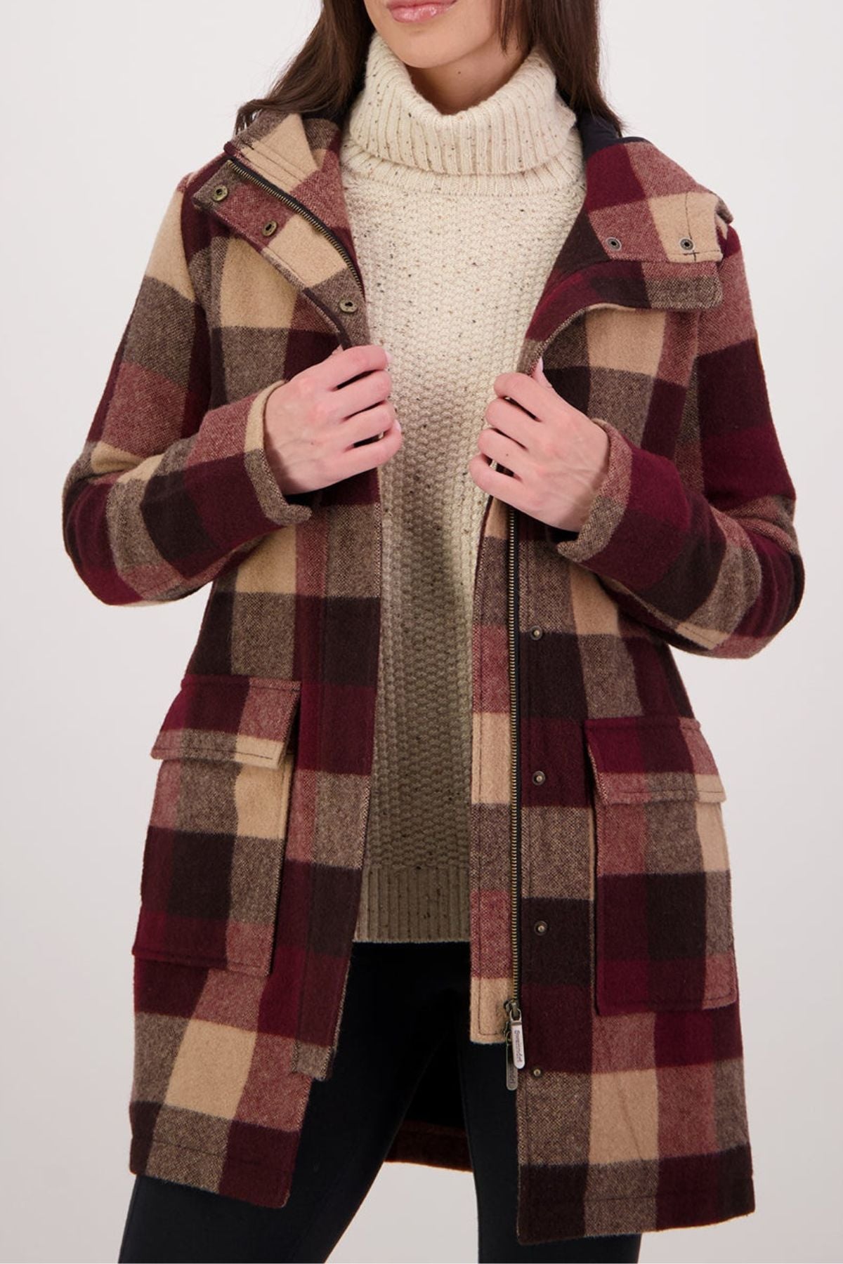 A woman in a beige turtleneck and a Swanndri Annmore V2 Wool Overcoat—long, plaid in burgundy, brown, and tan with large front pockets and a zipper—stands against a light background.