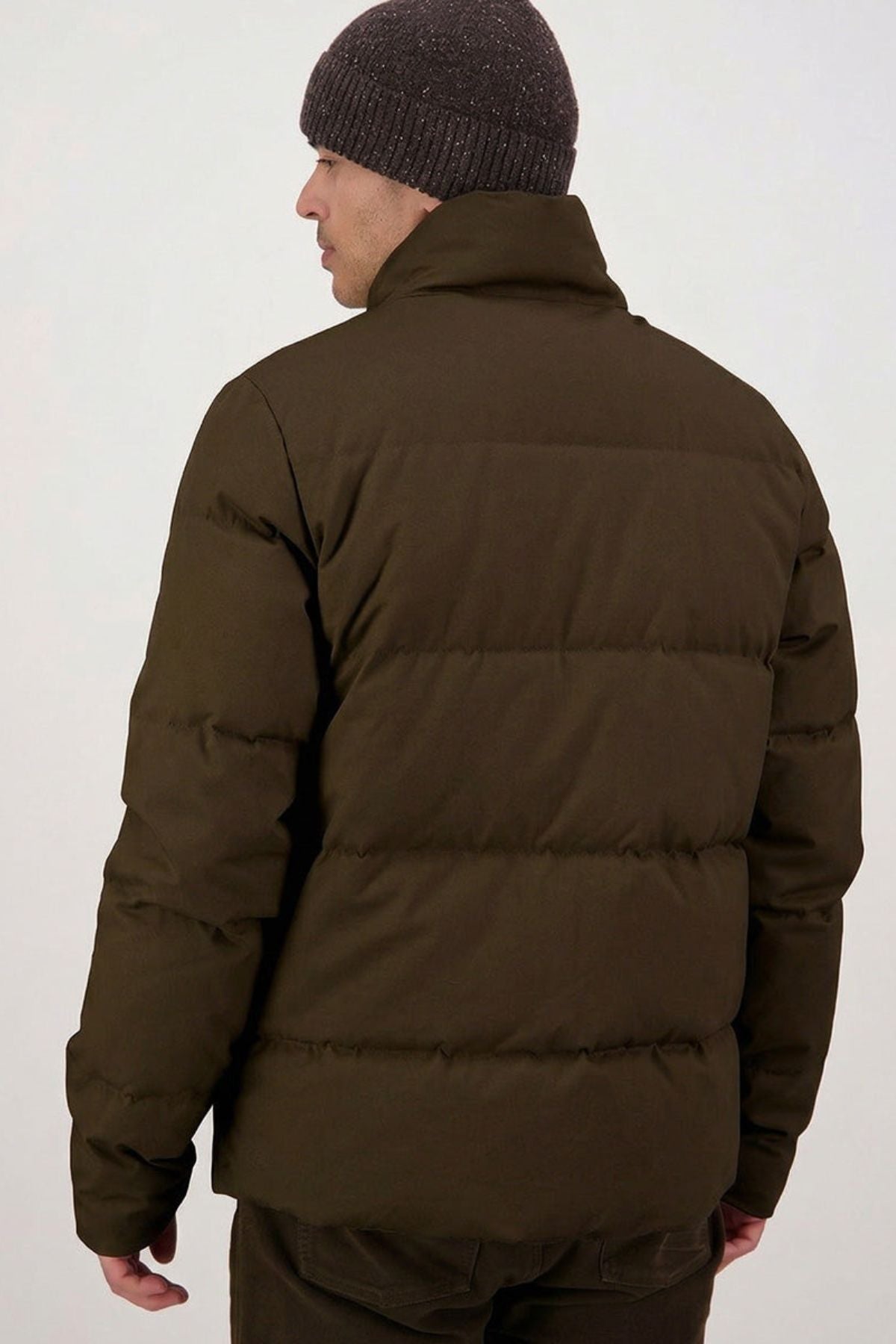 Wearing a dark brown Swanndri Nimbus Down Jacket and a gray knitted beanie, a person stands with their back to the camera against a plain, light background.