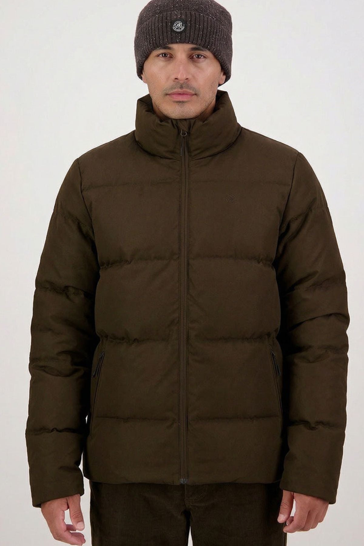 A man wears the Swanndri Nimbus Down Jacket in brown and a dark knit beanie, standing facing forward against a light plain background with a neutral expression and relaxed hands at his sides.