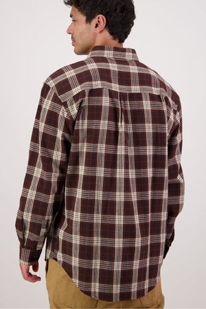 A man stands with his back to the camera in a brown, white, and red plaid Swanndri Barn Shirt with long sleeves and tan pants. The plain, light background highlights this ideal winter layering shirt.