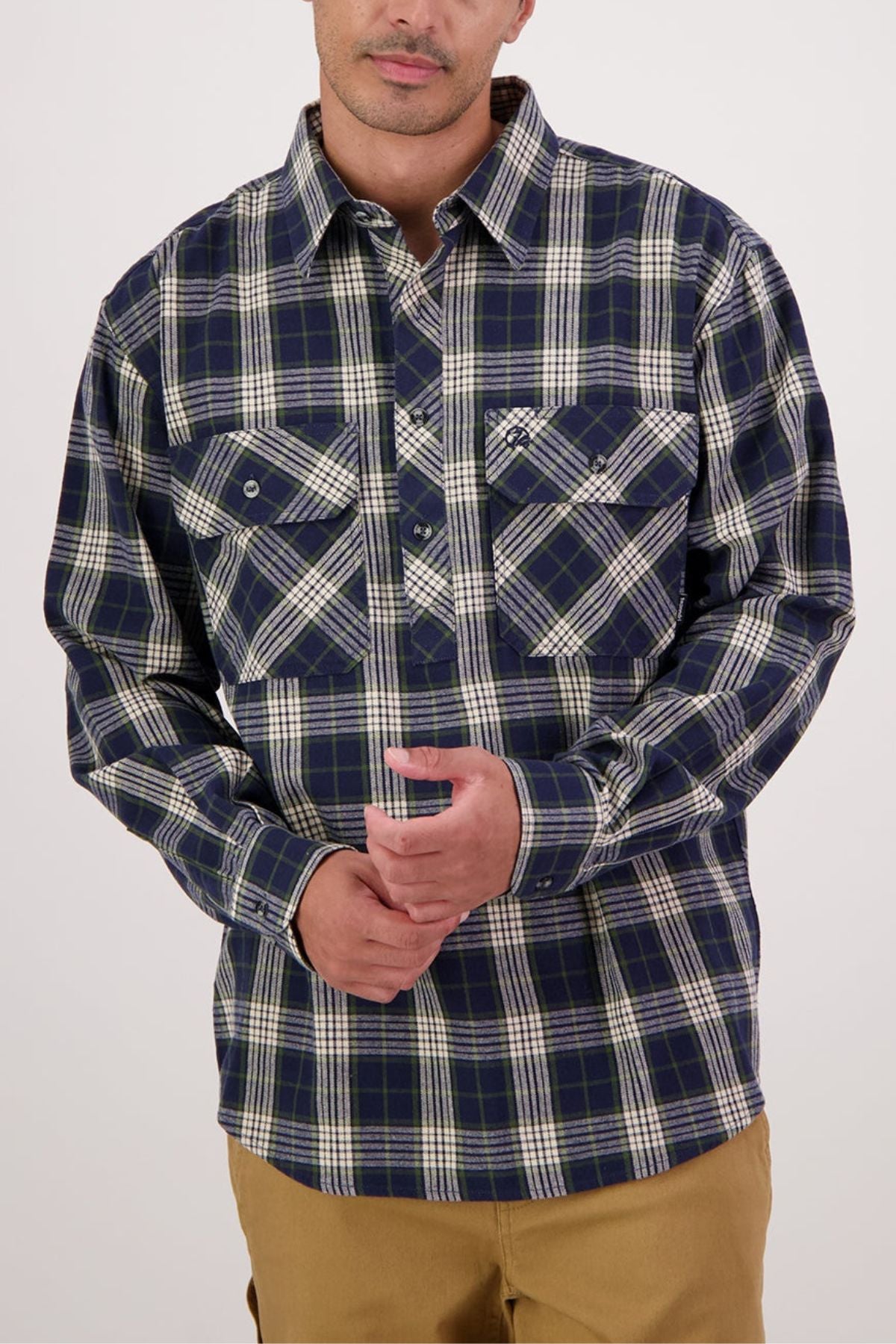 A person wears a Swanndri Barn Shirt in navy blue and white plaid with front pockets, ideal for winter layering, paired with tan pants. They stand with hands lightly clasped against a plain, light-colored background.