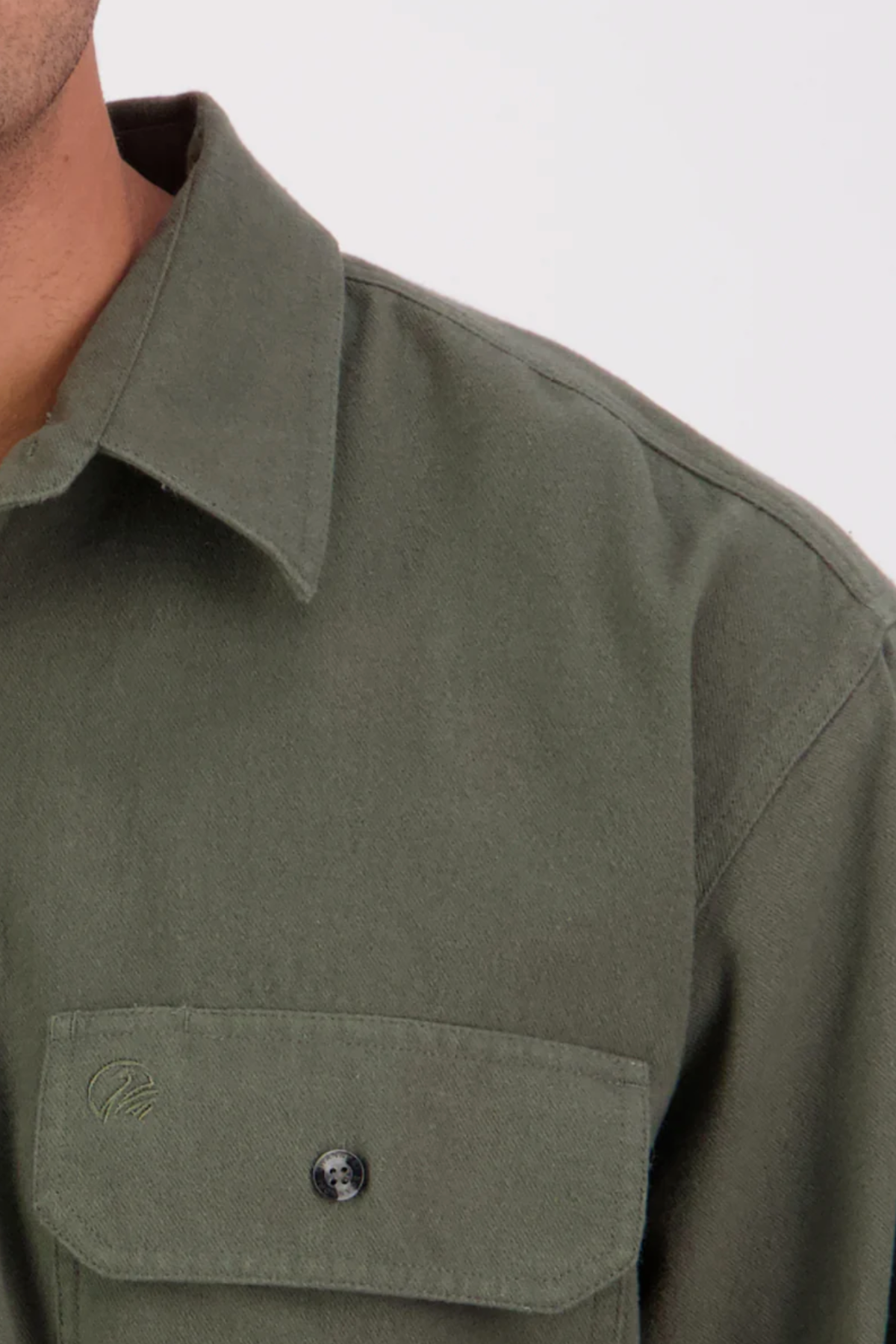 Close-up of the upper half of a person wearing the Swanndri Barn Shirt, with a collar, front pocket, and relaxed fit visible in olive green against a plain background. Only part of their shoulder, collar, and chest are shown.
