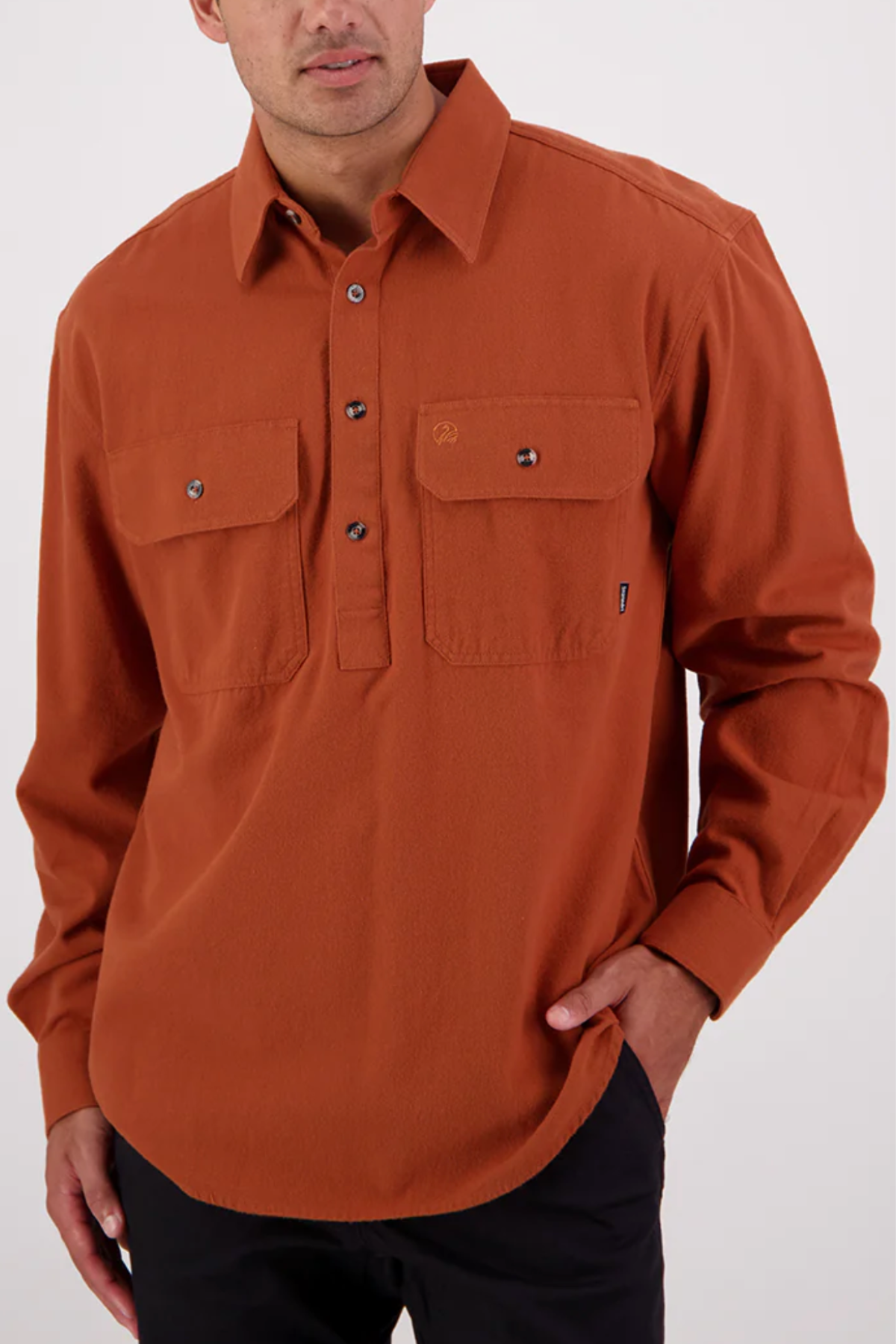 A man stands against a plain light background in a rust-orange Swanndri Barn Shirt—relaxed fit, 100% cotton, with two chest pockets—paired with black pants and one hand in his pocket.