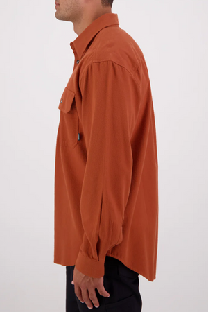 A man stands in profile wearing a relaxed-fit, long-sleeve rust-orange Swanndri Barn Shirt with a chest pocket, made from 100% cotton, paired with dark pants against a plain light background.