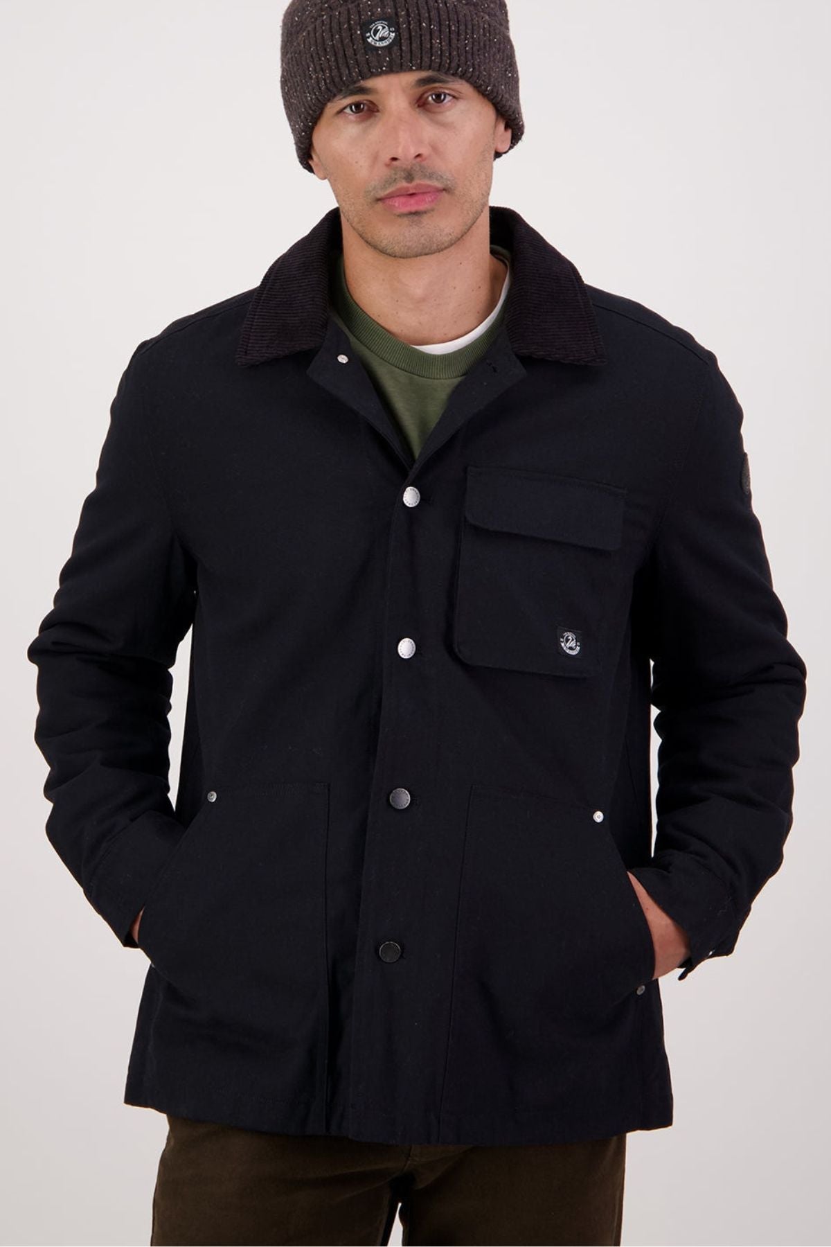 A man stands with his hands in the pockets of a Swanndri Barrytown V3 Canvas Jacket, paired with a gray knit beanie, green shirt underneath, and dark pants against a plain background.