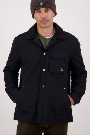 A man stands with his hands in the pockets of a Swanndri Barrytown V3 Canvas Jacket, paired with a gray knit beanie, green shirt underneath, and dark pants against a plain background.
