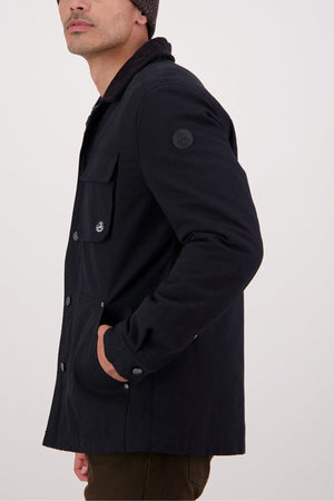A man wearing the Swanndri Barrytown V3 Canvas Jacket in black, brown pants, and a dark beanie stands sideways with one hand in his pocket against a plain light background.