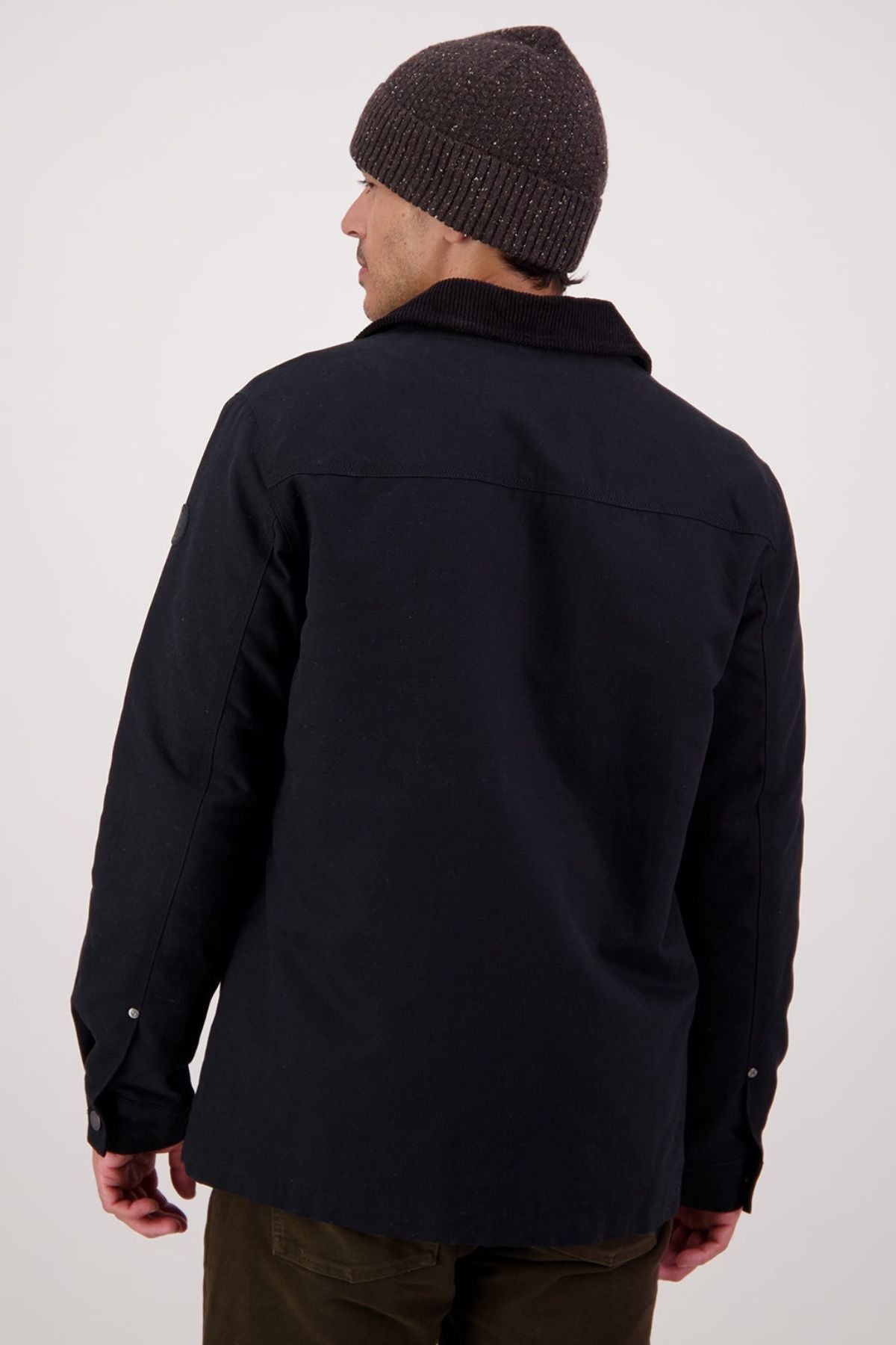 A person in a dark knit beanie and a Swanndri Barrytown V3 Canvas Jacket stands facing away from the camera against a plain, light background.