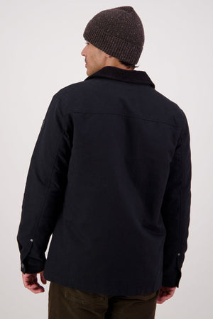 A person in a dark knit beanie and a Swanndri Barrytown V3 Canvas Jacket stands facing away from the camera against a plain, light background.