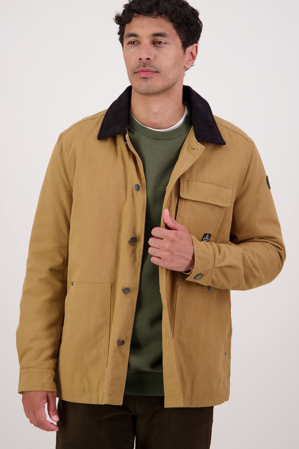 A man with short dark hair, wearing a Swanndri Barrytown V3 Canvas Jacket with a black collar over a green sweatshirt, stands facing forward against a light background, holding one side of his jacket.