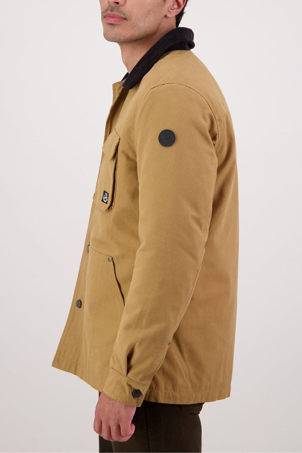 A man in a Swanndri Barrytown V3 Canvas Jacket with a black collar stands sideways against a plain, light background. The cotton canvas jacket features flap chest pockets and an upper arm patch.