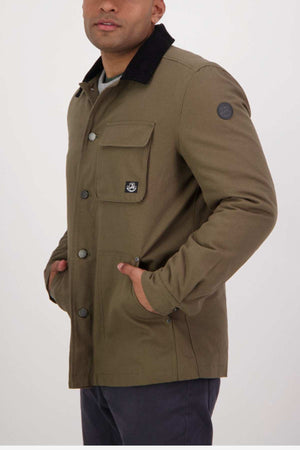 A person in an olive Swanndri Barrytown v3 Canvas Work Jacket with a black collar and button closure stands with hands in pockets. Known for its durability, it features a chest pocket and logo patch on the left sleeve against a plain white background.