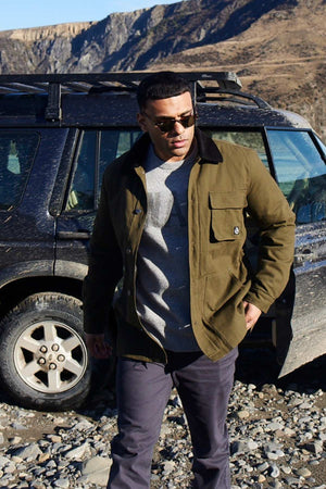 A man wearing a Swanndri Barrytown v3 Canvas Work Jacket, gray sweater, and dark pants stands before a muddy SUV on rocky terrain. The rugged backdrop with rocky hills and blue sky highlights an ideal outdoor setting for testing this durable workwear.