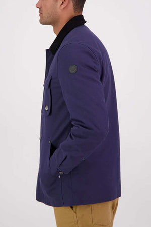 A man in a blue Swanndri Barrytown v3 Canvas Work Jacket with a black collar and beige pants stands in profile, one hand in his pocket, against a plain light background.