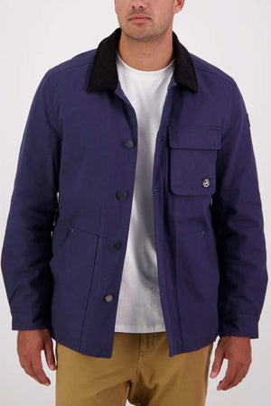 A man wearing a Swanndri Barrytown v3 Canvas Work Jacket in purple with a black collar, over a white t-shirt and tan pants, stands against a plain background.
