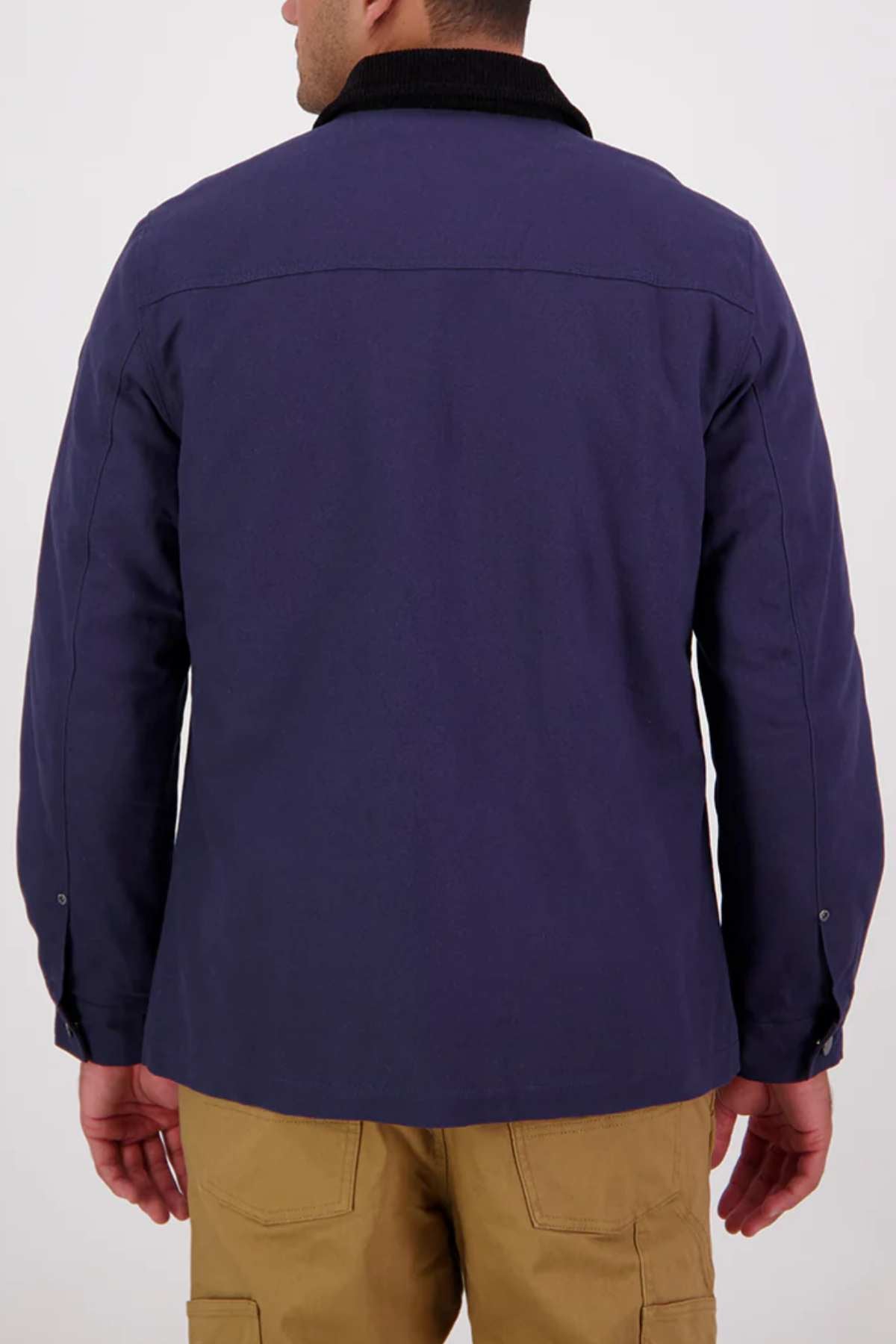 A man stands with his back to the camera, wearing a dark blue Swanndri Barrytown v3 Canvas Work Jacket with a black collar and tan pants. The cotton canvas material contrasts with the plain, light-colored background.