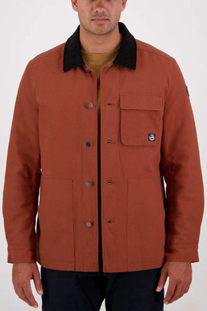 A man wears the Swanndri Barrytown v3 Canvas Work Jacket in rust, featuring black buttons, a black collar, reinforced stitching, a chest pocket with flap, and black pants against a plain light background.