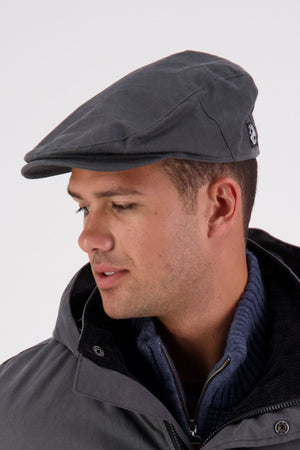 A man wearing a Swanndri Birmingham Oilskin Flat Cap, a dark zip-up sweater, and a gray jacket looks to his left against a plain light background.